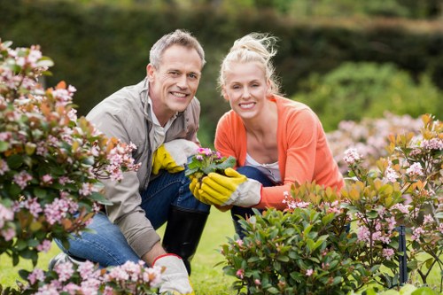 A man and woman are outdoors in a well-maintained garden in Sanderstead, surrounded by blooming pink and white flowering bushes, with lush green grass and neatly trimmed hedge borders in the background. They are both smiling at the camera, wearing gardening gloves, and appear to be engaged in plant maintenance or pruning activities. The scene is illuminated by natural daylight, suggesting fair weather with soft sunlight. The garden features a mix of flowering shrubs and grassy lawn, bordered by flower beds and small trees, reflecting careful landscaping and outdoor upkeep. This image exemplifies professional garden maintenance and hedge trimming services provided by Hedge Trimming Sanderstead, highlighting a tidy, vibrant, and accessible outdoor space designed for enjoyment and aesthetic appeal.
