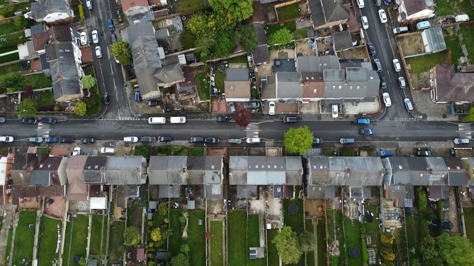 An aerial view of a residential street showcasing well-maintained gardens and lawns behind rows of terraced houses with tiled roofs. The gardens feature neatly trimmed lawns, planted borders with vari